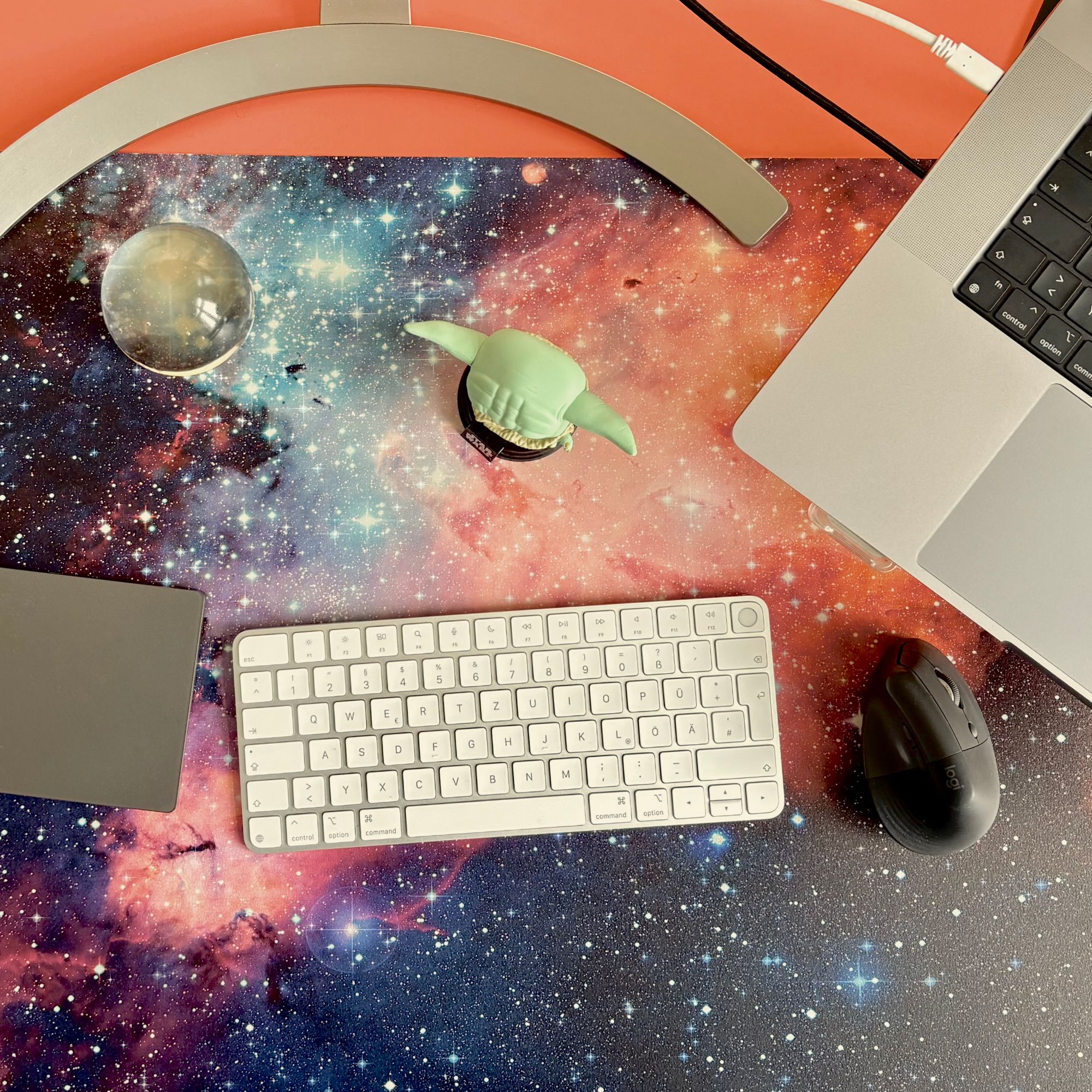 A modern desk with a galactic desk mat, laptop, keyboard, trackpad, mouse, and a glass sphere.
